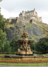 The ornate fountain in Princess Street Gardens, Edinburgh, with the historic Edinburgh Castle perched on the hill behind it. The fountain features intricate sculptures and flowing water, set against a backdrop of lush greenery and the castle’s dramatic stone walls.