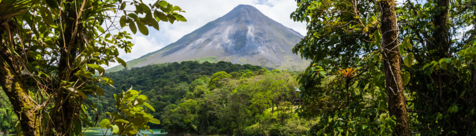 : A panoramic view of Arenal Volcano in Costa Rica, surrounded by lush tropical rainforest. The towering volcano rises above the dense greenery, framed by vibrant foliage under a partly cloudy sky.
