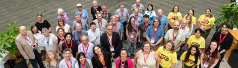 A diverse group of University of Guelph alumni and current students gather for a group photo during an alumni event. They are smiling and wearing red lanyards with name tags, while some are in yellow t-shirts with a celebratory logo. The group is standing together in a spacious indoor area with greenery in the background.
