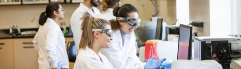 Two students wearing lab coats and safety goggles work together at laboratory equipment while other students work in the background.