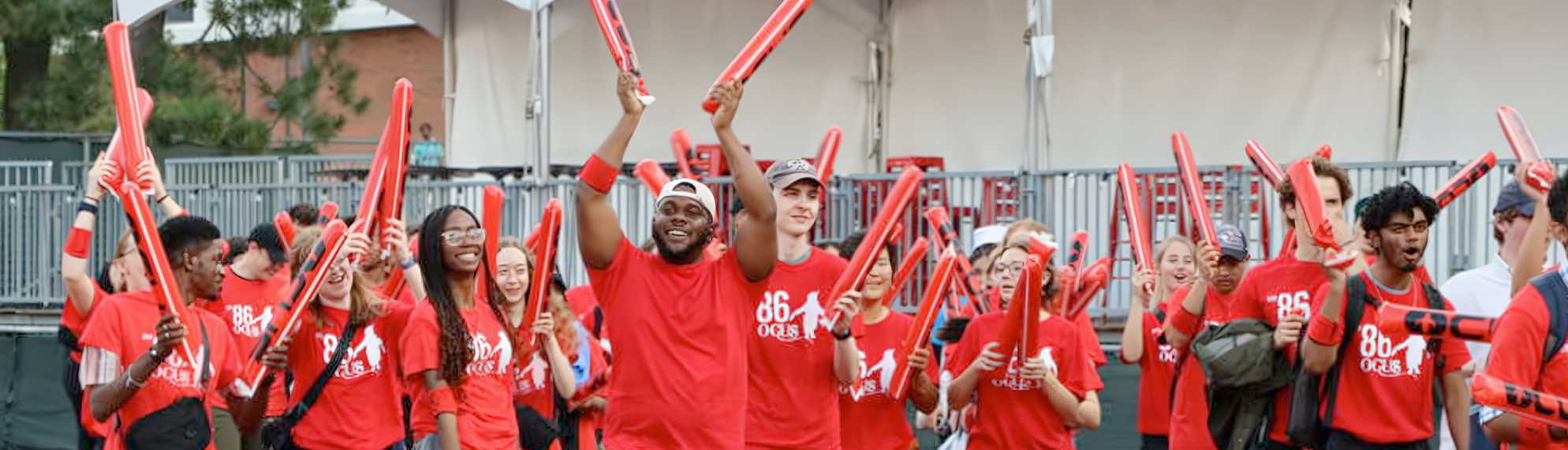 A large group of diverse young people wearing matching red shirts and wristbands enthusiastically raising red inflatable cheering sticks outdoors, appearing to celebrate or support an event.