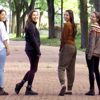 A group of Indigenous students walking together on a tree-lined campus pathway, smiling and looking back towards the camera, representing community and inclusivity at the University.