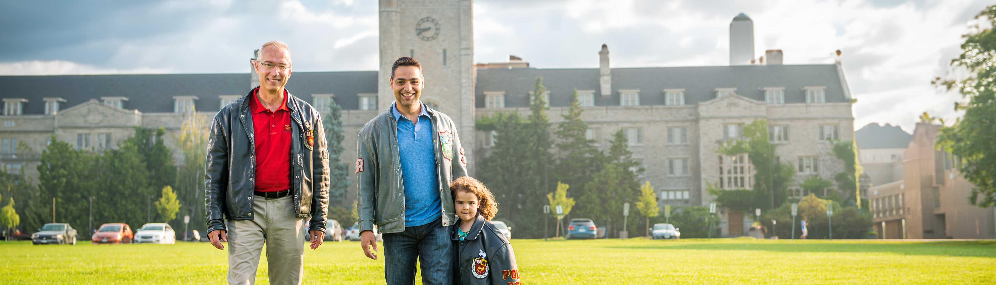 Two men and a young child wearing University of Guelph jackets standing on a grassy area in front of a historic building on the University of Guelph campus.