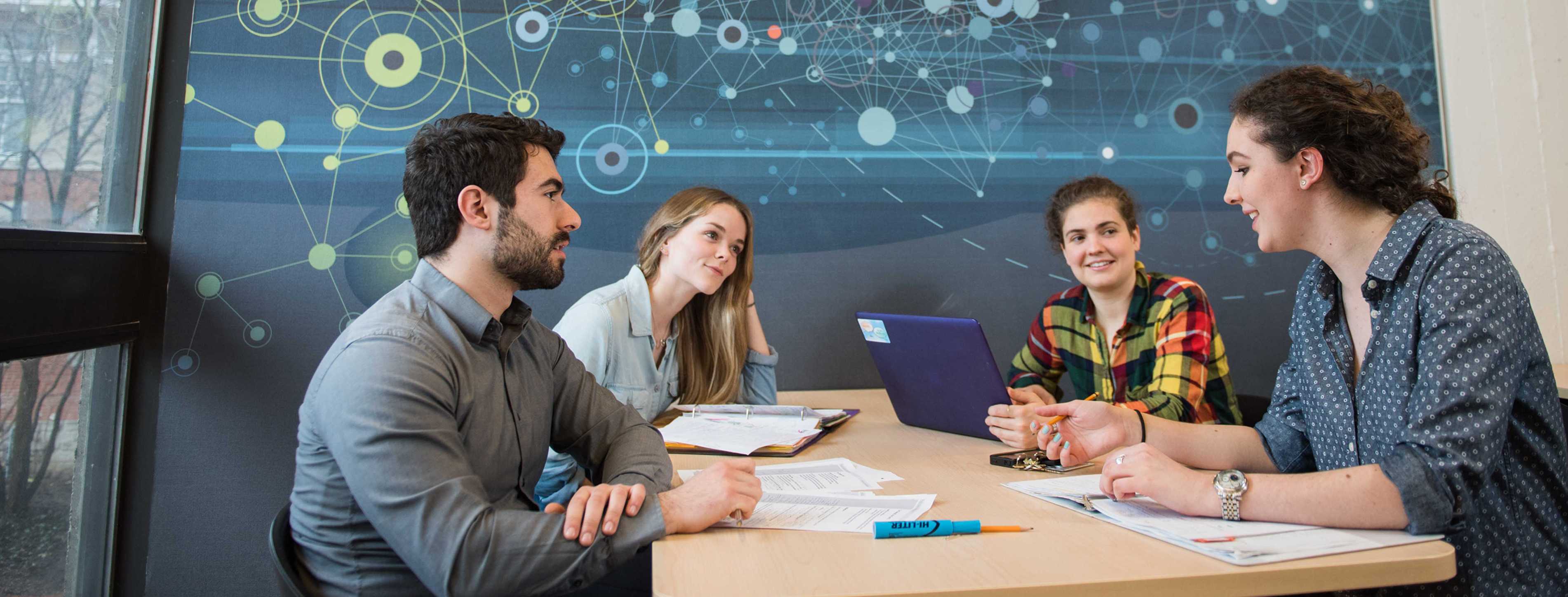 Four University of Guelph students sitting around a table during a study session. They are engaged in a discussion with notebooks and a laptop on the table. The background features a large window and a wall with an abstract scientific design.