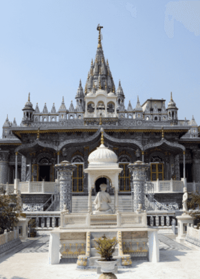The ornate Jain temple in Surat, India, with intricate carvings and stunning architectural details. The temple’s grand design is highlighted by its towering spires and the peaceful statue of a seated feature in the foreground, set against a clear blue sky.