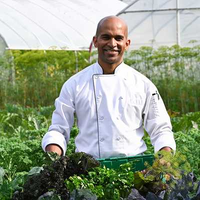A smiling chef wearing a white chef's coat stands in the middle of a lush green vegetable garden, holding a green crate filled with fresh leafy greens. In the background, there are greenhouses and tall plants under natural daylight.