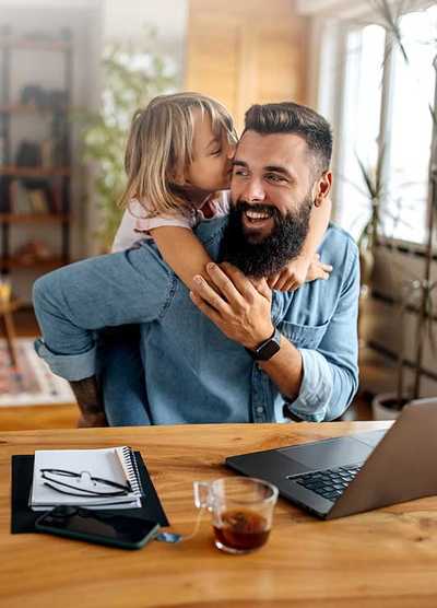 A young daughter kissing her bearded father on the cheek as he smiles, sitting at a desk with a laptop in a home office setting.