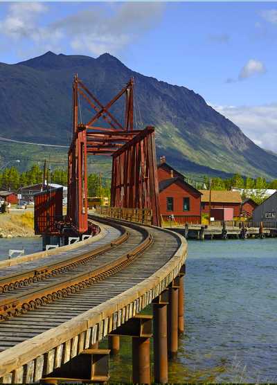 A historic red railway bridge in Carcross, Yukon, Canada, with mountainous terrain in the background and wooden tracks curving over the water.