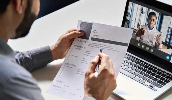 A person holding a CV in hand while attending an online job interview via video call on a laptop. The laptop screen shows a man in professional attire speaking during the virtual interview.